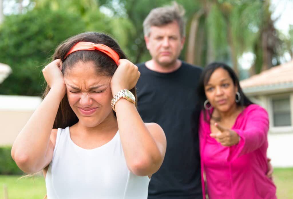 Teenage girl holding her head with very upset parents in the background